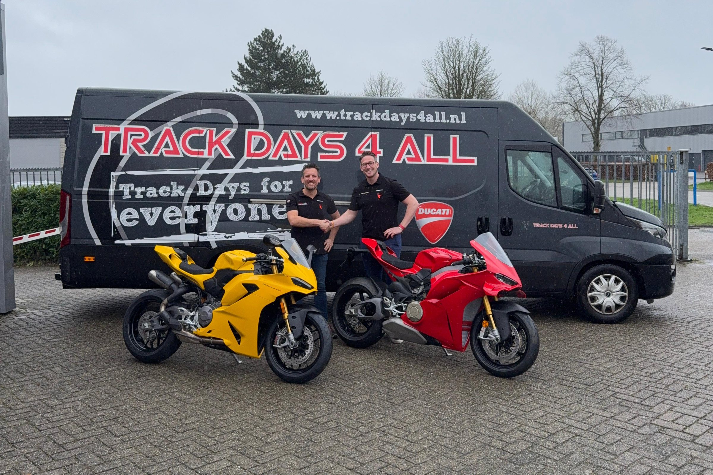 Twee mannen geven elkaar een hand voor de Trackdays4all-bus, met een gele en een rode Ducati Panigale ervoor